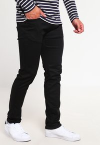 Pantalon noir à coupe slim, fabriqué dans un tissu lisse avec des poches avant. Porté avec un haut à rayures à manches longues et des baskets blanches.