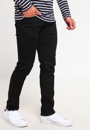 Pantalon noir à coupe slim, fabriqué dans un tissu lisse avec des poches avant. Porté avec un haut à rayures à manches longues et des baskets blanches.