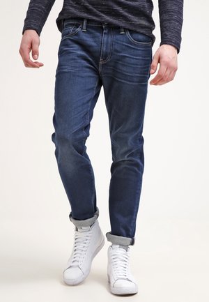 Homme portant un jean bleu foncé retroussé et des baskets blanches montantes, debout devant un fond clair uni.