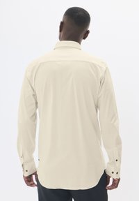 Világos bézs színű, hosszú ujjú ing, gallérral, sima textúrával, gombos mandzsettákkal és ívelt alsó szegéllyel. A hátsó nézet varrási részleteket mutat.