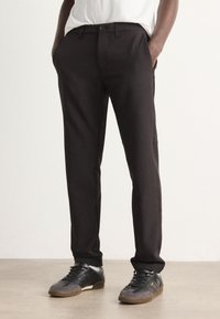 Black, tailored pants with a smooth texture, featuring belt loops, front pockets, and a slim fit. Paired with black sneakers.