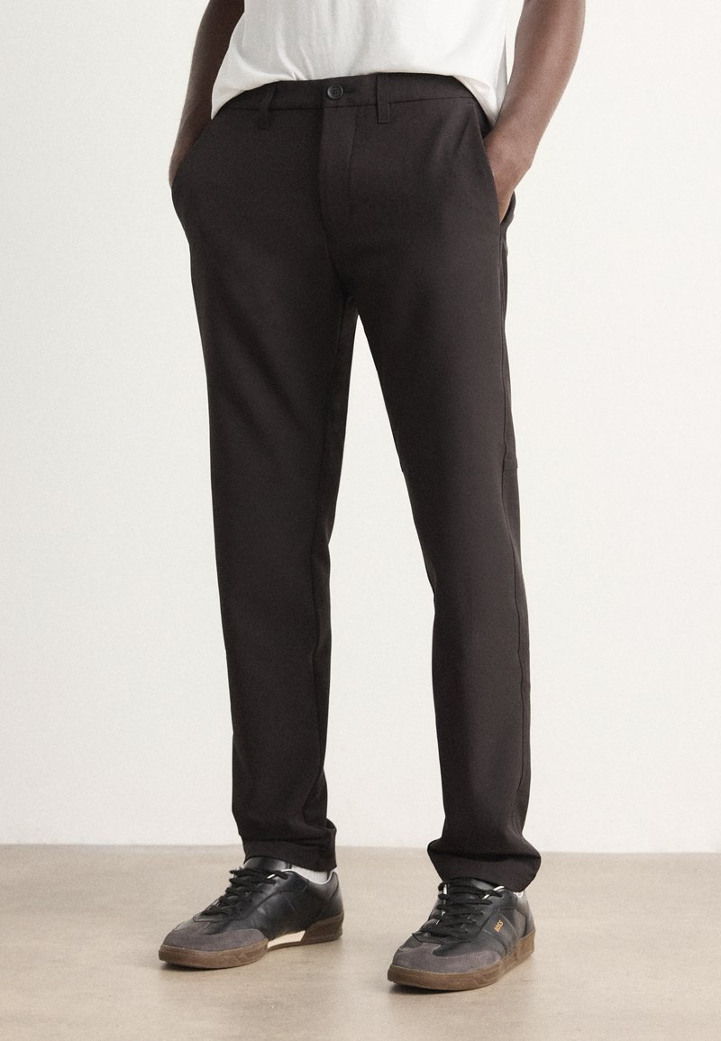 Black, tailored pants with a smooth texture, featuring belt loops, front pockets, and a slim fit. Paired with black sneakers.