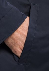 Granatowa kurtka zbliżenie na rękę w bocznej kieszeni; gładki materiał z widocznym stębnowaniem i prostym otworem kieszeni.