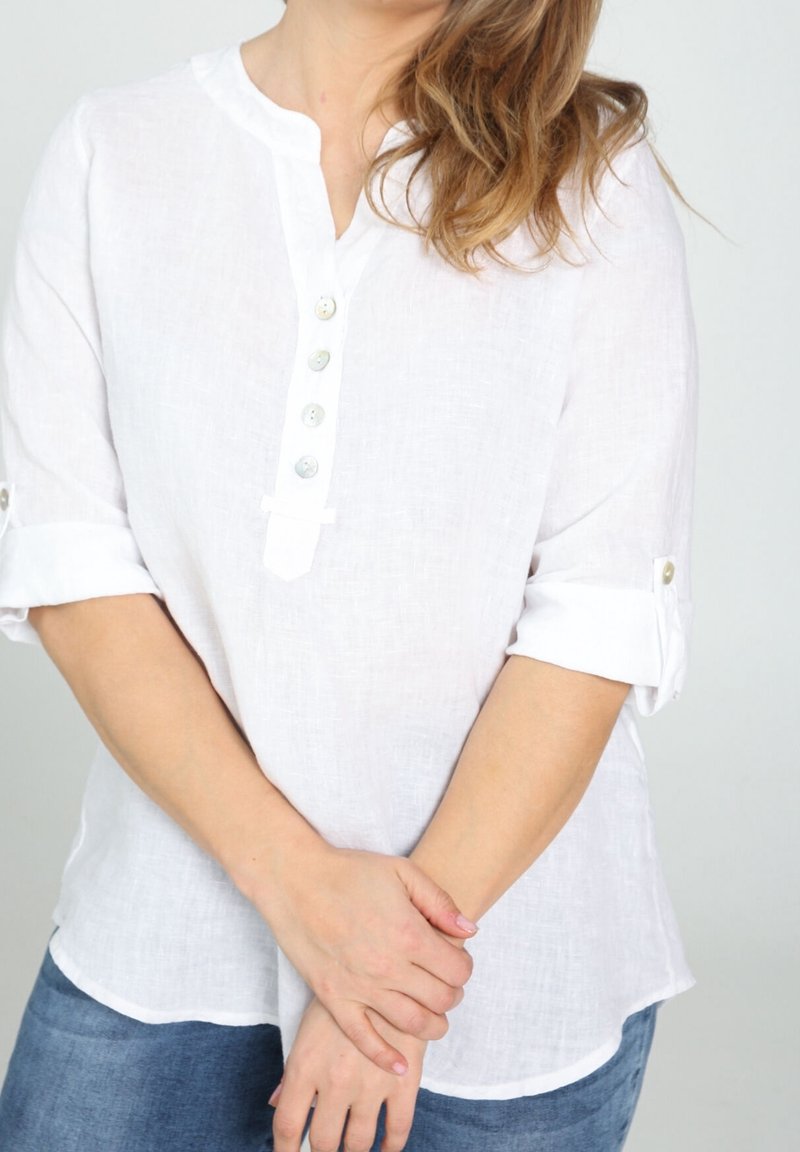 Blouse en lin blanc avec une patte de boutonnage à six boutons, manches retroussables avec des boutons décoratifs et un ourlet arrondi. Portée avec un jean en denim bleu.