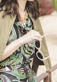 Abito a stampa tropicale verde con accenti rosa, giallo e nero; sovrapposto a una giacca verde oliva. Tiene occhiali da sole bianchi; accessori dorati.