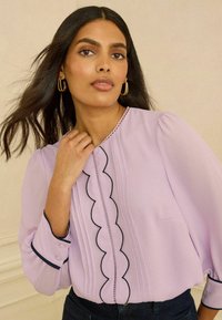 Laventelin värinen pusero, joka on valmistettu kevyestä kankaasta. Siinä on kaarevat reunukset, laskokset sekä kontrastiväriset tummansiniset koristeet kauluksessa ja kalvosimissa.