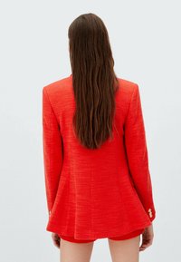Blazer rouge texturé avec un ourlet évasé, deux accents de boutons dorés sur les manches, et une coupe ajustée, porté sur un short, vu de derrière.