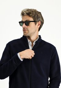 Granatowa polarowa kurtka z zamkiem, noszona na jasnoniebieskiej koszuli z kołnierzykiem; zestawiona z okularami w kolorze żółwia; gładka tekstura.