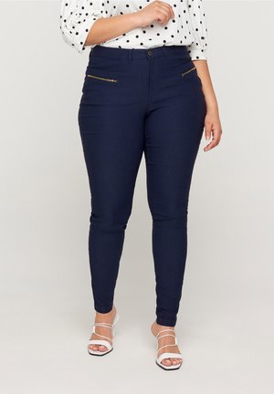 Frau trägt marineblaue, eng anliegende Hose mit vorderen Reißverschlusstaschen, eine weiße Bluse mit Punkten und weiße, rückenfreie Sandaletten mit Absatz.