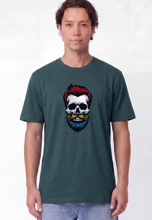 Homme portant un t-shirt vert foncé avec un graphique coloré de crâne avec cheveux rouges, moustache jaune et barbe bleue, debout sur fond blanc.