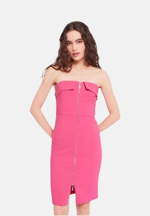 Vrouw met lang krullend haar die een felroze strapless jurk met een ritssluiting aan de voorkant draagt en staat met haar handen achter haar rug.