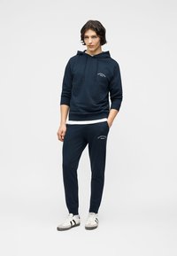 Νεαρός άνδρας στέκεται φορώντας καπελοφόρο φούτερ σε ναυτικό μπλε και αντίστοιχο παντελόνι φόρμας με λευκά αθλητικά παπούτσια, πάνω σε απλό φόντο.