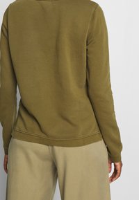 Persoon in een olijfgroene langemouwenshirt en beige broek, gezien van achteren tegen een effen witte achtergrond.