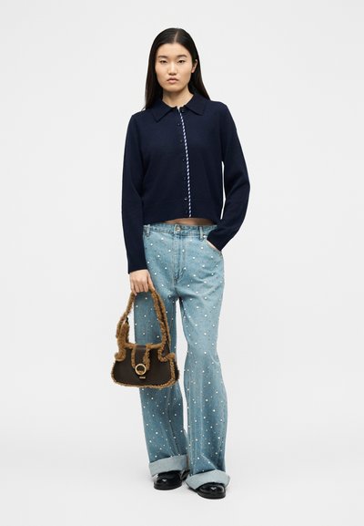 Γυναίκα φορώντας μπλε καφέ ζακέτα με κουμπιά, ανοιχτό μπλε τζιν με πουά, μαύρα παπούτσια, κρατώντας καφέ αφράτη τσάντα, σε απλό φόντο.