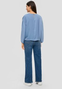Hellbluse mit langen Ärmeln, Rundhalsausschnitt und Knopfdetails auf der Rückseite, kombiniert mit blauen weit geschnittenen Jeans und weißen Turnschuhen.