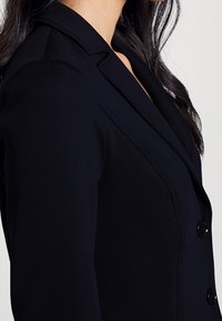 Blazer navy con design sartoriale, colletti a falda, spalle strutturate, texture liscia e due bottoni frontali.