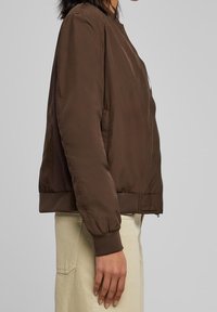 Chaqueta bombér marrón con una textura suave, puños acanalados y cierre de cremallera. Presenta bolsillos laterales y un diseño ligeramente recortado.