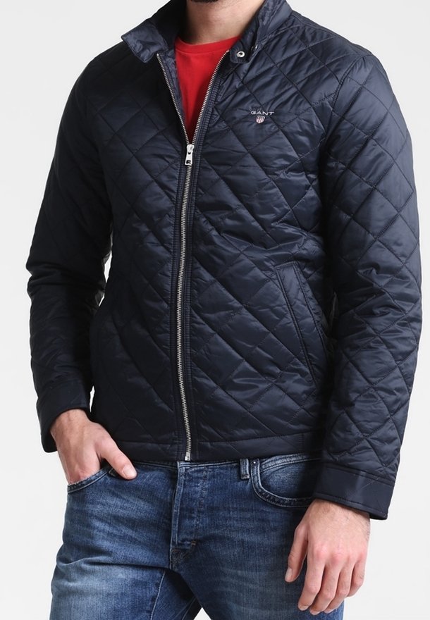 Veste matelassée marine avec fermeture éclair, col montant et détail de logo. Portée sur une chemise rouge et associée à un jean en denim.