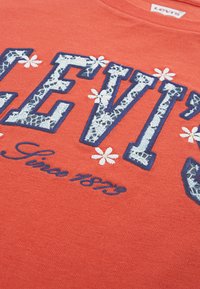 Κόκκινο βαμβακερό T-shirt με το λογότυπο «LEVI'S» σε ναυτικό μπλε, διακοσμημένο με floral μοτίβα, υφασμάτινη δαντέλα και την επιγραφή «Since 1873» με καλλιγραφικά γράμματα κάτω από αυτή.