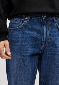 Vaqueros de mezclilla azul oscuro con detalles visibles de costuras, cinco bolsillos y cierre de botones. La tela parece suave con una ligera textura.