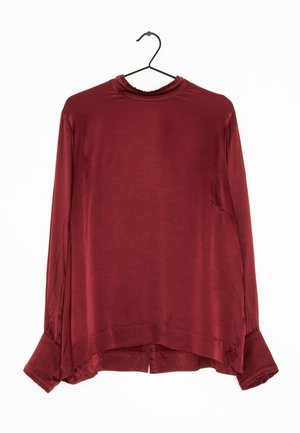 Blusa de satén burdeos de manga larga con cuello alto y puños abotonados colgada en una percha negra sobre un fondo blanco.