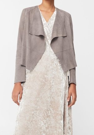 Femme portant une robe portefeuille beige clair texturée avec une veste grise douce drapée à ouverture devant, debout devant un fond uni.