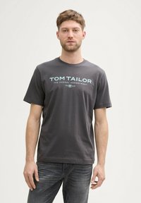Pilkas medvilninis marškinėliai trumpomis rankovėmis ir apvaliu kaklu. Centruotas šviesiai mėlynas logotipo tekstas skelbia „TOM TAILOR“ su sklandžiu paviršiumi.