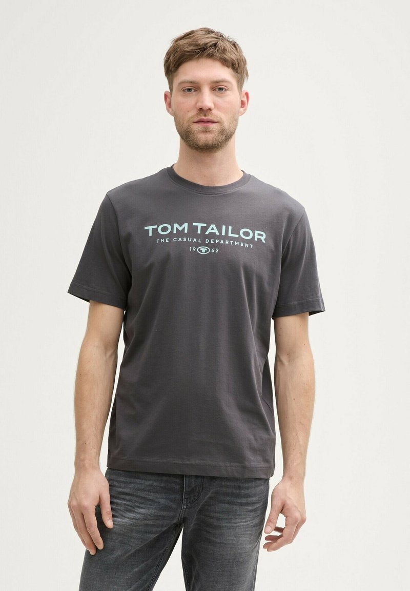 Pilkas medvilninis marškinėliai trumpomis rankovėmis ir apvaliu kaklu. Centruotas šviesiai mėlynas logotipo tekstas skelbia „TOM TAILOR“ su sklandžiu paviršiumi.