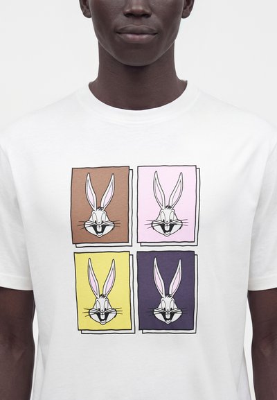 Άνδρας που φοράει λευκό T-shirt με τέσσερα καρτουνίστικα πρόσωπα κουνελιών μέσα σε καφέ, ροζ, κίτρινους και σκούρους μωβ τετράγωνους στο μπροστινό μέρος.