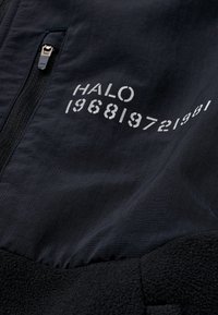 Czarna kurtka z fakturowanej tkaniny, z dolną częścią z polaru oraz białym nadrukiem "HALO 196819721981" w górnej części.