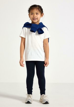 Ung pige smilende, iført hvid t-shirt, mørke leggings, sneakers, med en blå sweater bundet om skuldrene, står på ensfarvet baggrund.