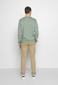 Felpa verde chiaro con orlo e polsini a costina, abbinata a pantaloni beige. Il modello è di spalle, mostrando una vista casual. Design pulito, colori neutri.