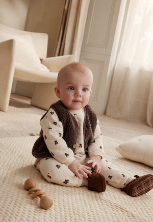 Bambino seduto su un tappeto beige che indossa un gilet marrone e una tutina crema con stampe di orsi, che gioca con un sonaglio di legno vicino a una sedia e un cuscino crema.