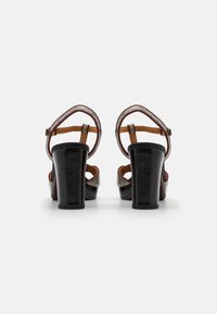 Black heeled sandals with a textured finish, featuring a patterned strap and a thick, solid heel. Adjustable ankle strap included.