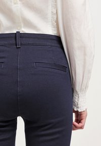 Pantalones azul marino con una textura suave, que presentan un corte ajustado, bolsillos traseros y un lazo para el cinturón, combinados con una blusa de color claro.