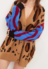 Brun leopardmönstrad cardigan med oversized passform, stora knappar och långärmade i lysande blå och röd mönster, bärs av en person.