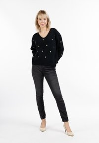 Pulover negru cu modele de inimi aurii, design în formă de V, asociat cu blugi skinny negri și pantofi cu toc bej, pe un fundal uniform.