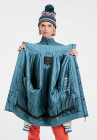 Tyrkysová nepremokavá bunda so zipsami a gombíkmi, vybavená izolačnou podšívkou a viacerými vreckami. Nosí sa s pruhovaným čiapkou.