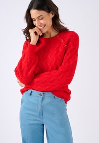 Donna sorridente in un luminoso maglione rosso a trecce con bottoni sulla spalla, che indossa jeans a vita alta azzurri, su sfondo bianco.