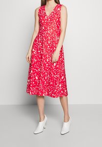 Mujer vestida con un vestido sin mangas rojo con patrones de salpicaduras blancas y botas de tacón alto blancas hasta el tobillo, de pie contra una pared lisa.
