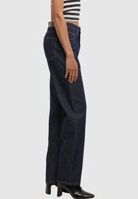 Person, die dunkelblaue Jeans mit weitem Bein, schwarze Absatzstiefel und ein schwarz-weiß gestreiftes Crop-Top trägt, seitlich vor einem einfarbigen Hintergrund steht.