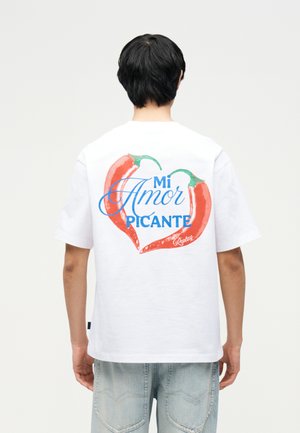 Osoba otočená chrbtom má na bielom tričku dva červené chilli papričky vytvárajúce srdce a modrý nápis „Mi Amor Picante“ na chrbte.