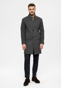 Grauer Wollmantel mit hohem Kragen, einreihigem Design, zwei Vorder Taschen, kombiniert mit dunkler Hose und braunen Wildlederschuhen.