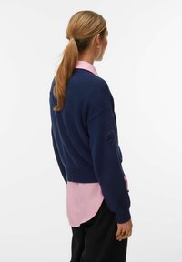 Femme aux cheveux châtain clair attachés, portant un pull bleu marine par-dessus une chemise rose et un pantalon noir, de profil devant un fond uni.
