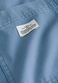 Mėlynas audinys su tekstūruota paviršiumi, turintis baltą etiketę, ant kurios yra užrašyta "STANDARD ISSUE" ir priežiūros instrukcijos, siuvamos krašte.