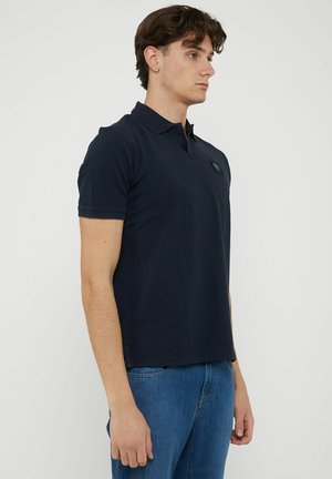 Giovane uomo con polo blu navy e jeans blu, in piedi di profilo su uno sfondo bianco uniforme.