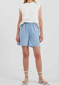Top sans manches blanc à côtes avec col rond, associé à un short en taille élastique bleu clair. Le modèle porte des sandales blanches à lanières.