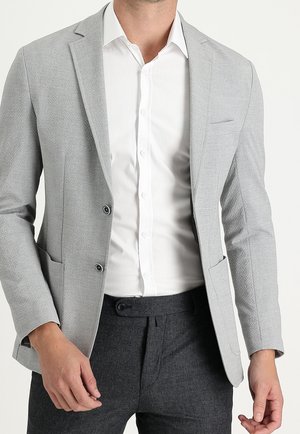 Hellgrauer Blazer aus strukturiertem Stoff, mit Reverskragen und zwei schwarzen Knöpfen. Getragen über einem weißen Hemd und dunklen Hosen.