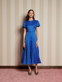 Vestido azul texturizado com mangas bufantes, comprimento até à meia panturrilha e um corpete ajustado. Combinado com sapatos de salto de padrão de leopardo contra um fundo riscado.