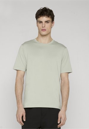 Ung mann med kort krøllete hår som har på seg en lys grønn kortermet t-skjorte med rund hals og svarte bukser, står foran en ensfarget bakgrunn.